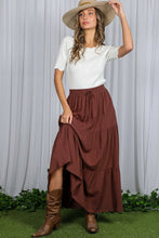 Load image into Gallery viewer, Woman wearing a white top, brown tiered skirt, and brown boots against a white curtain background.