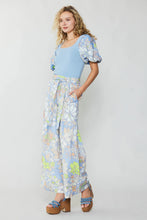 Load image into Gallery viewer, The immaculately dressed model adorns a stylish blue top paired with wide-leg pants boasting a retro floral print.
