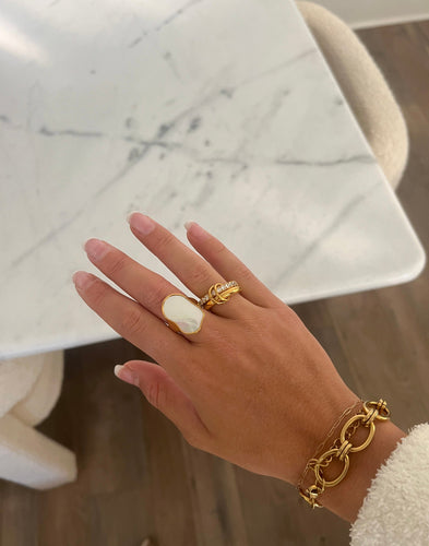 A gold ring featuring an irregular, lustrous white mother-of-pearl stone set atop, displayed on a smooth gray surface, exuding elegance and luxury.