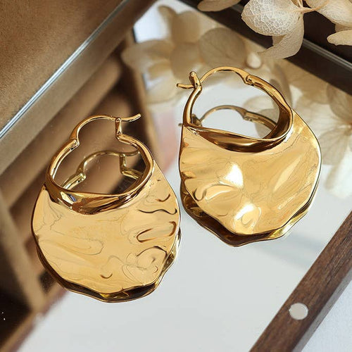 A pair of gold-plated, wavy design hoop earrings on a reflective surface.